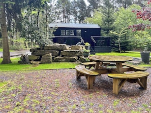 A wooden picnic table with benches, a stone wall, and a building with a metal roof in a forested area. - Shepherds Hut - UK52010 (Harrogate)