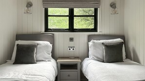 A bedroom with two single beds, a nightstand, a window with a view of greenery, and wall-mounted lights. - Shepherds Hut - UK52010 (Harrogate)