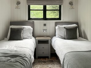 A bedroom with two single beds, a nightstand, a window with a view of greenery, and wall-mounted lights.