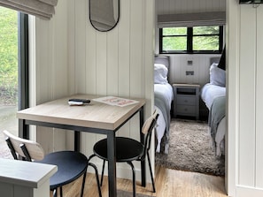 A room with a wooden table, two chairs, a bed with white bedding, a nightstand, and a window with a view of greenery. - Shepherds Hut - UK52010 (Harrogate)
