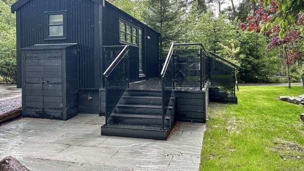 A black metal building with a staircase leading to a glass door entrance, surrounded by greenery. - Shepherds Hut - UK52010 (Harrogate)