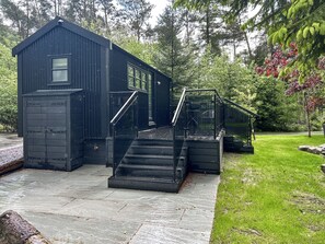 A black metal building with a staircase leading to a glass door entrance, surrounded by greenery.
