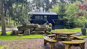 A wooden picnic table with benches, a stone wall, and a building with a metal roof in a forested area. - Shepherds Hut - UK52010 (Harrogate)