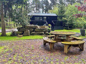A wooden picnic table with benches, a stone wall, and a building with a metal roof in a forested area.