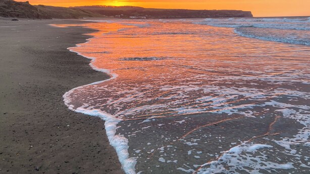 Una playa con olas, un atardecer y colinas a lo lejos.