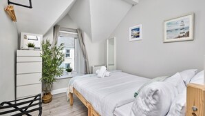 Quarto com teto inclinado, uma cama com roupa de cama branca, uma mesa de cabeceira de madeira, uma planta em vaso e uma janela com cortinas.