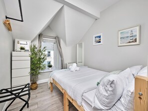 A bedroom with a sloped ceiling, a bed with white bedding, a wooden nightstand, a potted plant, and a window with curtains. - No.3 Princess Blue Apartments (Portrush)
