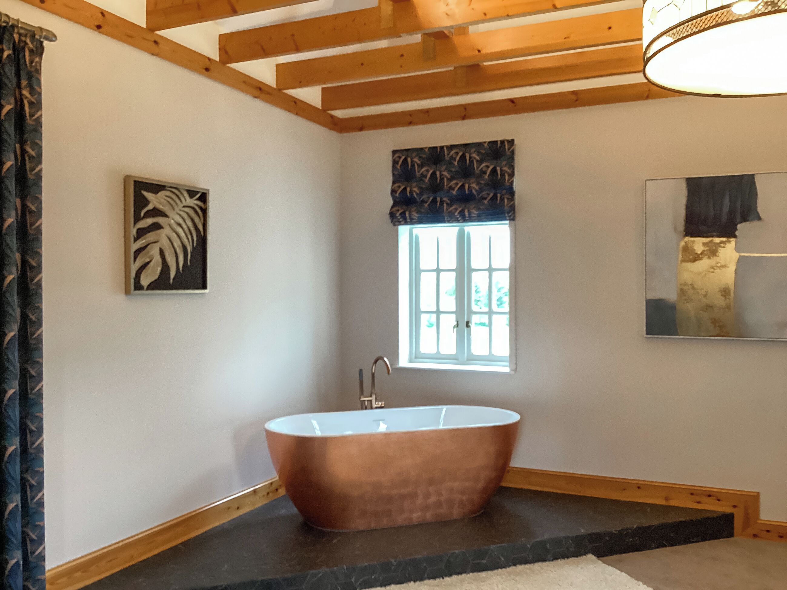 A bathroom with a freestanding bathtub, a window with a view, and a painting on the wall.