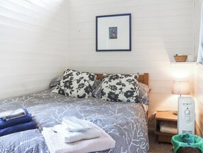 A neatly made bed with floral-patterned pillows, a framed picture above it, and a bedside table with a lamp and a green trash bin.