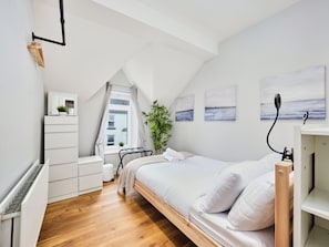 Ein Schlafzimmer mit einem hölzernen Bett, einer weißen Kommode, einem Heizkörper, einem Fenster mit Vorhängen und einer Pflanze.