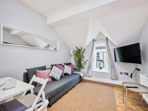 Ein modernes Wohnzimmer mit grauem Sofa, Flachbildfernseher, kleinem Tisch und einer Pflanze.