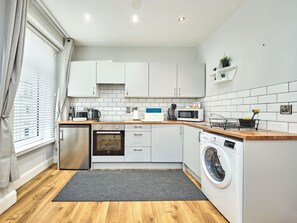 Eine kompakte Küche mit weißen Schränken, einer Mikrowelle, einer Kaffeemaschine, einer Waschmaschine und einem kleinen Fenster mit Jalousien.