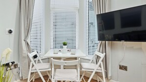 Quarto com mesa branca, duas cadeiras, televisão e janela com persianas.