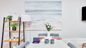 Un moderno comedor con una mesa blanca, sillas tapizadas de gris, un jarrón blanco con flores, una estantería con plantas y un cuadro abstracto de gran tamaño en la pared.