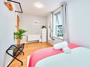 Ein Schlafzimmer mit Bett, Heizkörper, einem Fenster mit Vorhängen, einer Pflanze und einer weißen Kommode.