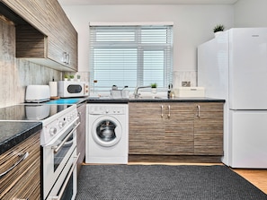 A modern kitchen with a built-in washer, oven, and microwave, wooden cabinetry, and a large window with blinds. - No.1 Princess Yellow Apartments - UK52052 (Portrush)
