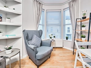 A modern living room with a grey armchair, a small table, and a white radiator. There are potted plants on shelves and a window with blinds. - No.2 Princess Yellow Apartments - UK52053 (Portrush)