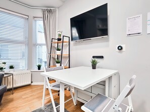 A modern room with a white table, chairs, a TV, a shelf with plants, and a window with blinds. - No.2 Princess Yellow Apartments - UK52053 (Portrush)