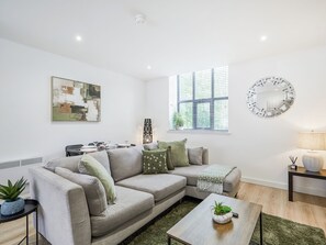 A modern living room with a sectional sofa, a coffee table, a side table with a lamp, a wall-mounted mirror, and a window with blinds. - Green Mill 3-UK52057 (Holmfirth)