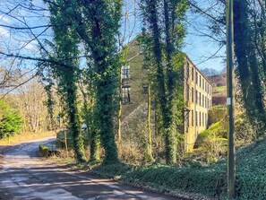 A stone building with ivy-covered walls, a paved road, and a clear sky. - Green Mill 3-UK52057 (Holmfirth)