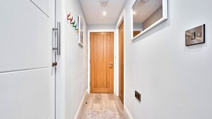 Un couloir aux murs blancs, une porte en bois, un miroir, un interrupteur et un tapis à motifs.