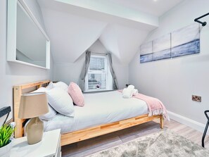 A bedroom with a sloped ceiling, a bed with a wooden frame, a bedside table with a lamp, a small potted plant, and a window with blinds. - No.3 Princess Yellow Apartments - UK52054 (Portrush)