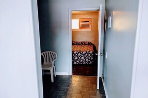 A narrow hallway leading to a bedroom with a bed, a chair, and a wall-mounted heater.