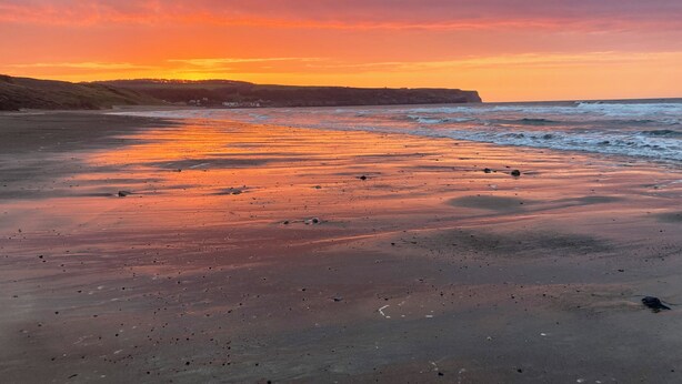 Uma praia com um pôr do sol, ondas e um penhasco ao longe.
