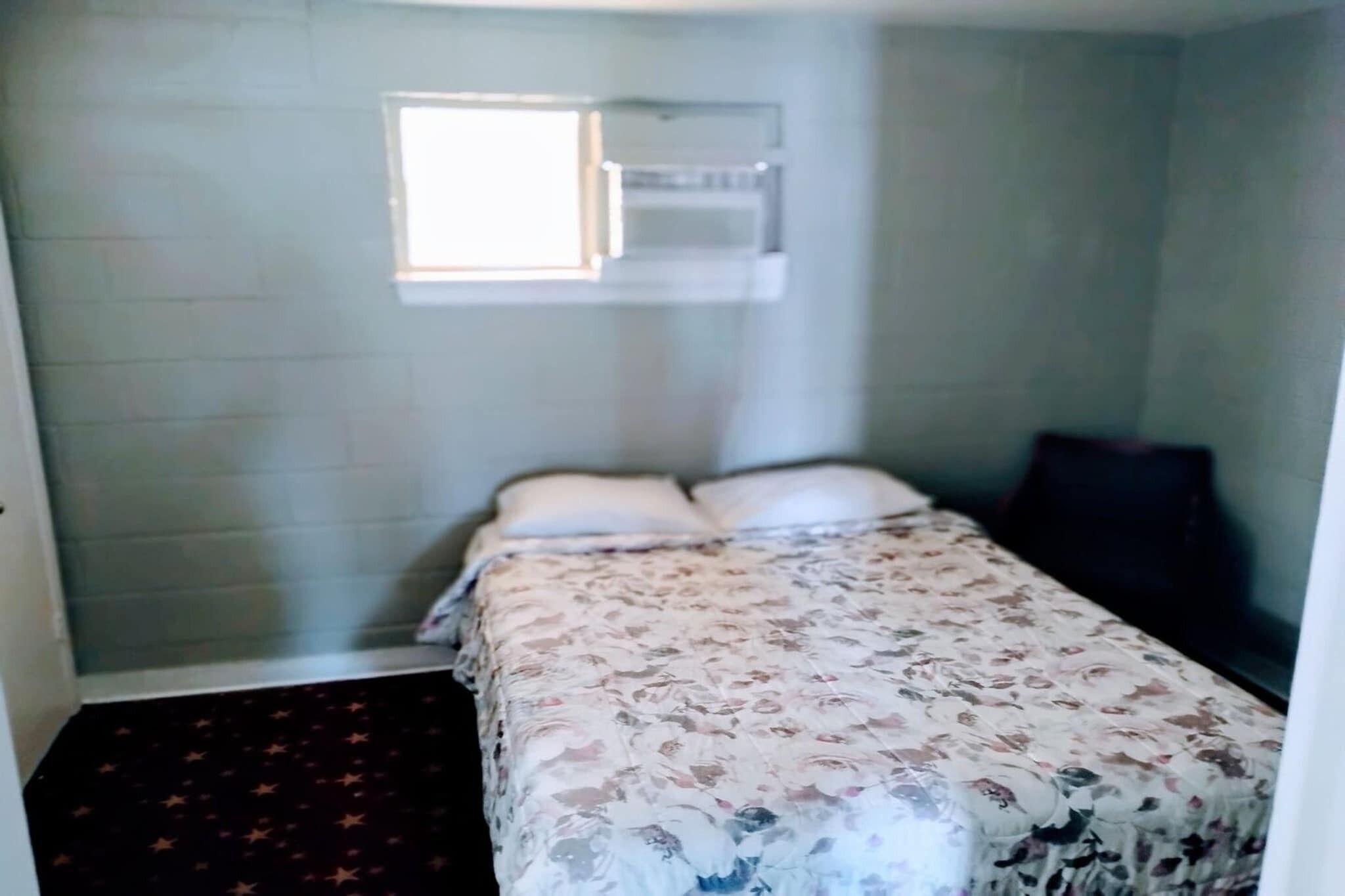 A small, single-bed room with a floral-patterned bedspread, a window with blinds, and a dark-colored chair.