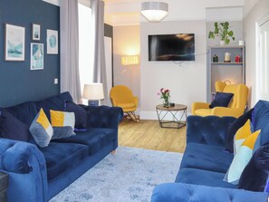 A living room with a blue sofa set, a yellow armchair, a round coffee table, and a flat-screen TV mounted on the wall. - 5 bedroom accommodation in Porthmadog (Porthmadog)