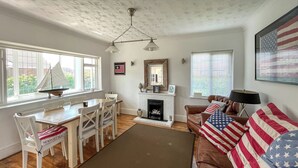 Ein Wohnzimmer mit Kamin, einem Esstisch mit Stühlen, einer Couch mit Kissen mit amerikanischer Flagge und einem großen Fenster mit Jalousien.