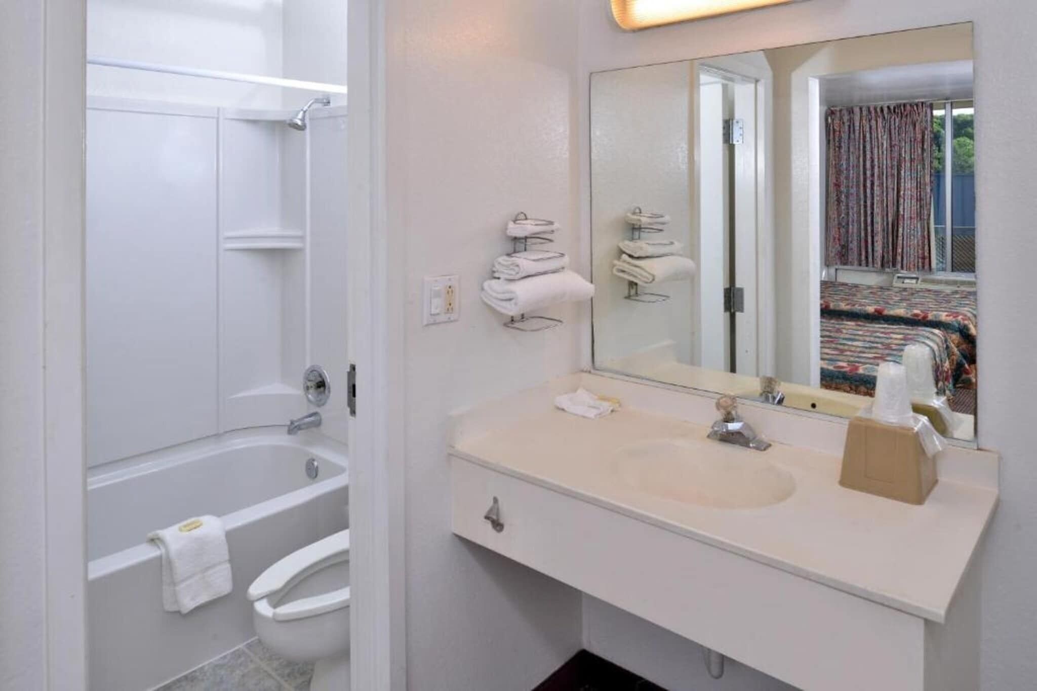A bathroom with a bathtub, toilet, sink, and a mirror reflecting a bed with curtains.