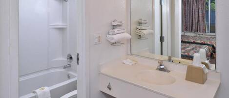 A bathroom with a bathtub, toilet, sink, and a mirror reflecting a bed with curtains.