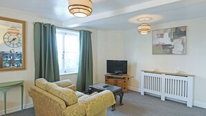 Een woonkamer met een bank, een televisie op een houten dressoir, een raam met gordijnen, een radiator en een wandklok.