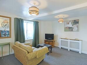 Een woonkamer met een bank, een televisie op een houten dressoir, een raam met gordijnen, een radiator en een wandklok.