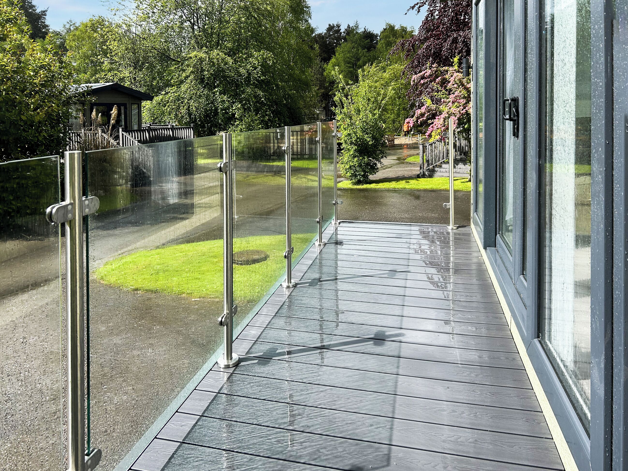 A covered outdoor area with a glass railing, a paved walkway, and a view of a park with trees and a building in the background.