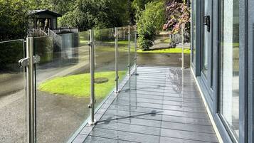A covered outdoor area with a glass railing, a paved walkway, and a view of a park with trees and a building in the background.