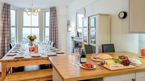 A dining area with a long wooden table set for a meal, a bottle of champagne, and a variety of fruits and cheeses. - Glan Y Mor - UK52092 (Criccieth)