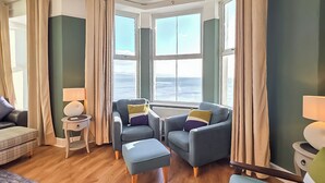 A room with a bed, two armchairs, a small table, and a view of the sea through large windows. - Glan Y Mor - UK52092 (Criccieth)