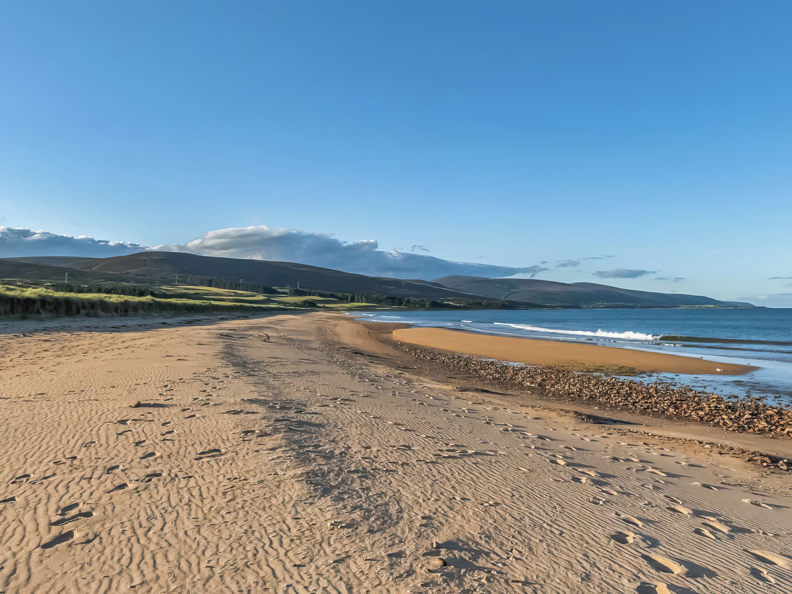 A beach with a clear sky, a sandy path leading to the water, and distant hills.