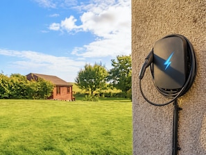 A grassy field with a house and trees in the background, and a wall-mounted electric charging station. - 1 bedroom accommodation in Brora (Brora)