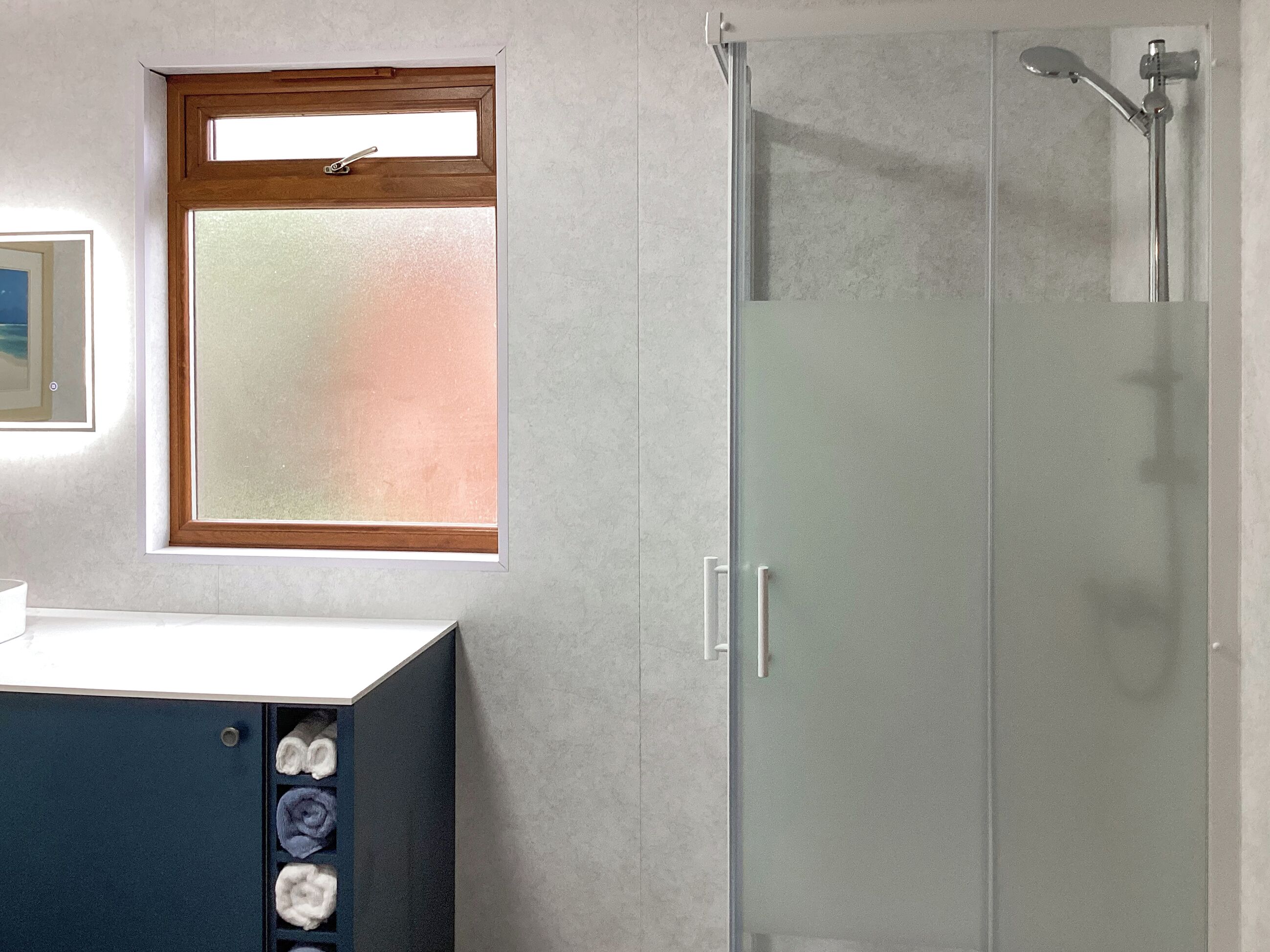 A bathroom with a glass shower enclosure, a wooden-framed window, and a white sink with towels.