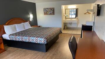 A hotel room with a bed, a desk, a chair, a bathroom with a mirror, and a wall-mounted TV.