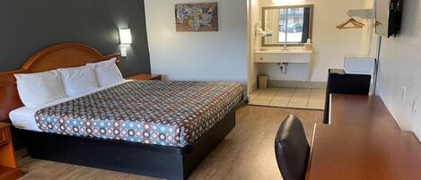 A hotel room with a bed, a desk, a chair, a bathroom with a mirror, and a wall-mounted TV.