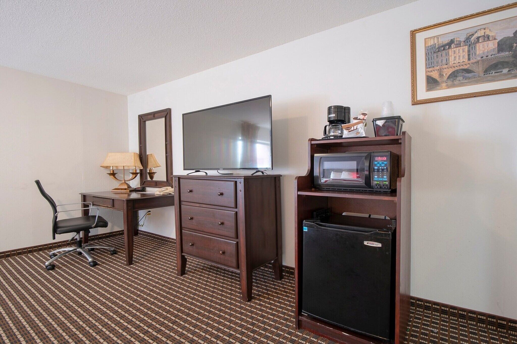 A hotel room with a desk, a television, a microwave, a refrigerator, a chair, and a framed picture on the wall.
