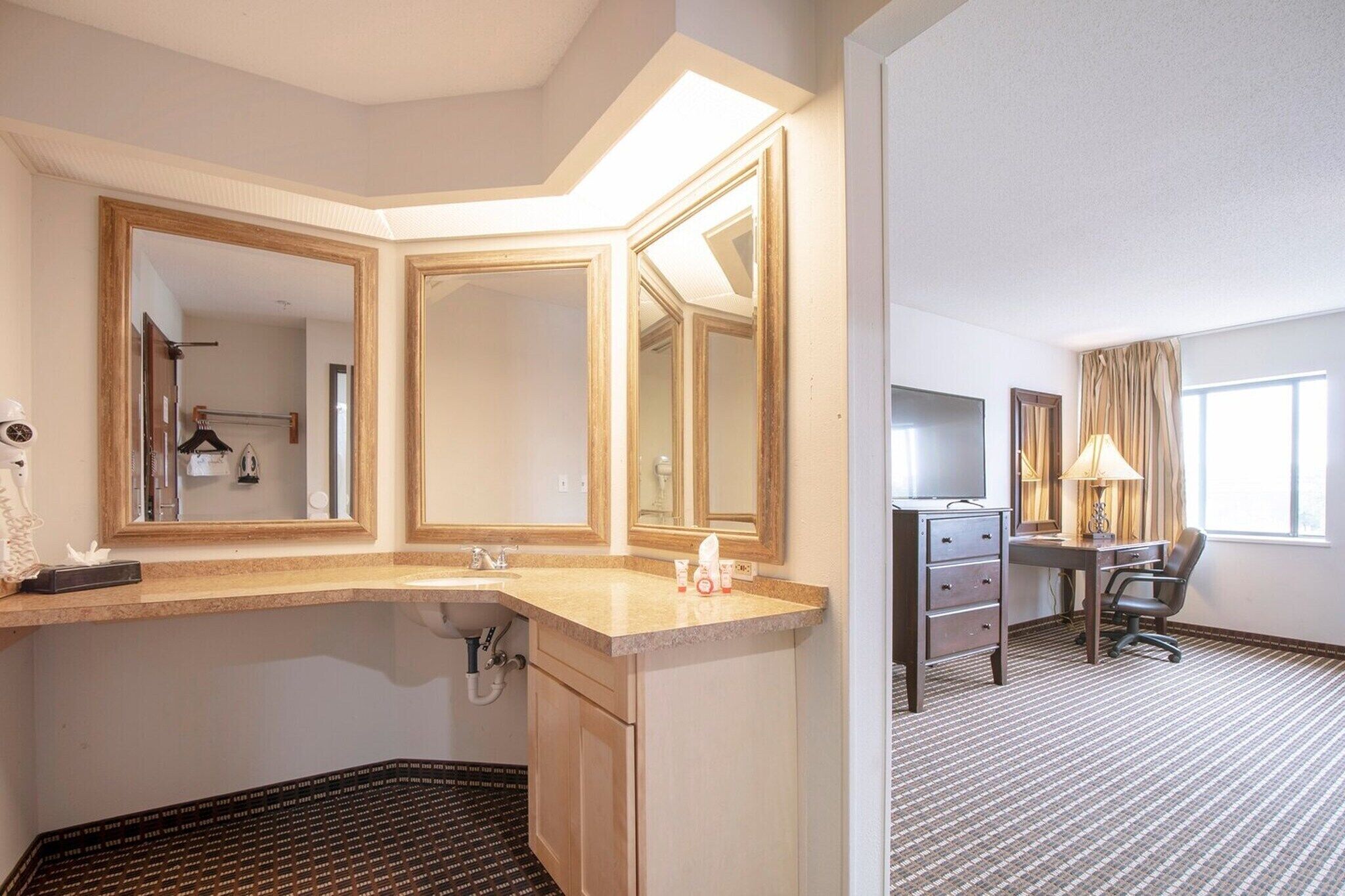 A hotel room with a bathroom, a desk, a chair, and a large window.