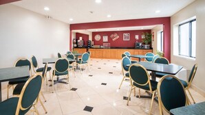 A dining area with tables and chairs, a kitchen in the background, and a clock on the wall.