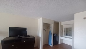 A room with a television on a dresser, a closet with a hanging rack, a small ironing board, and a bathroom with a mirror.