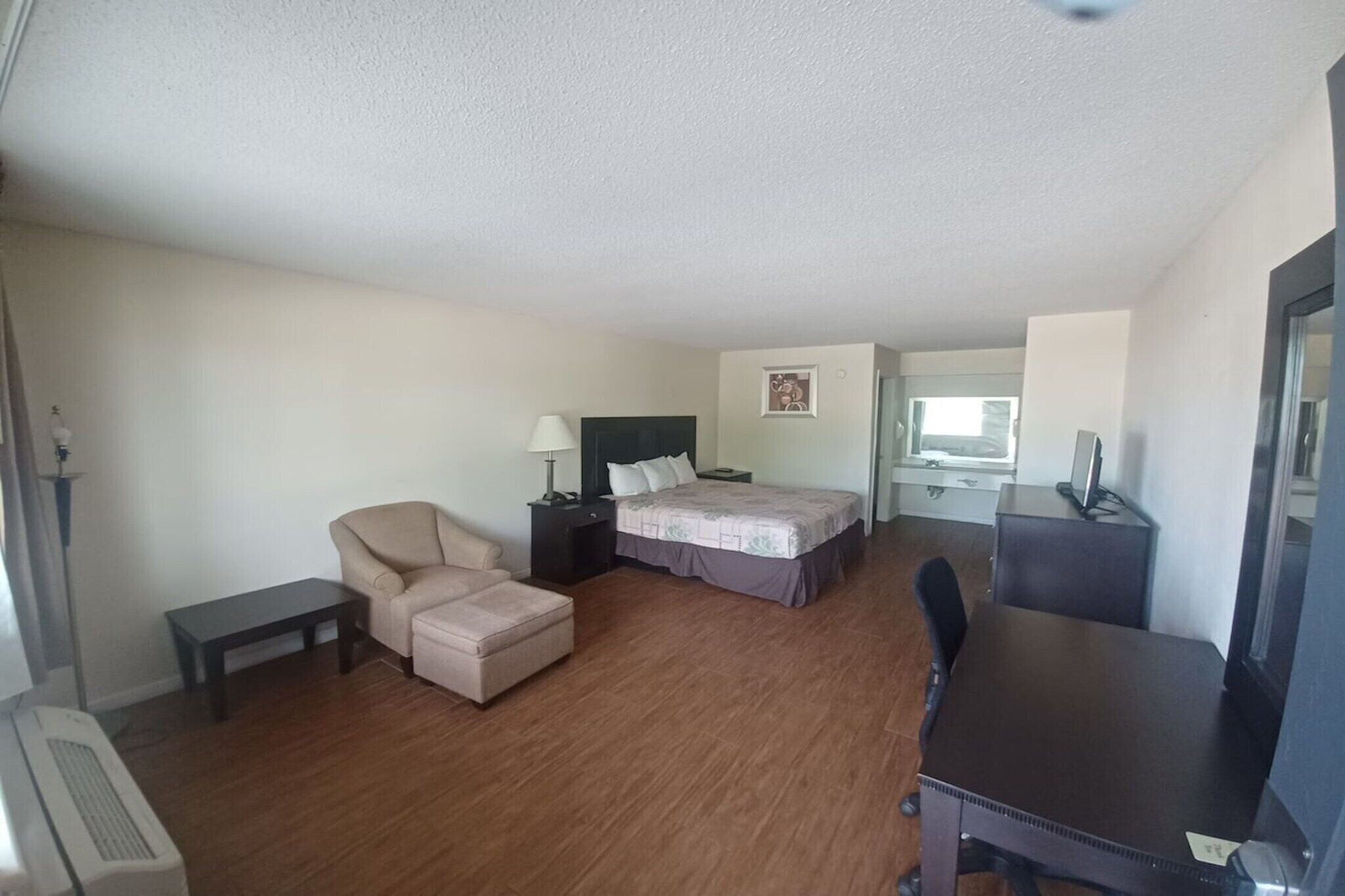A hotel room with a bed, a desk, a chair, a small table, and a bedside lamp.