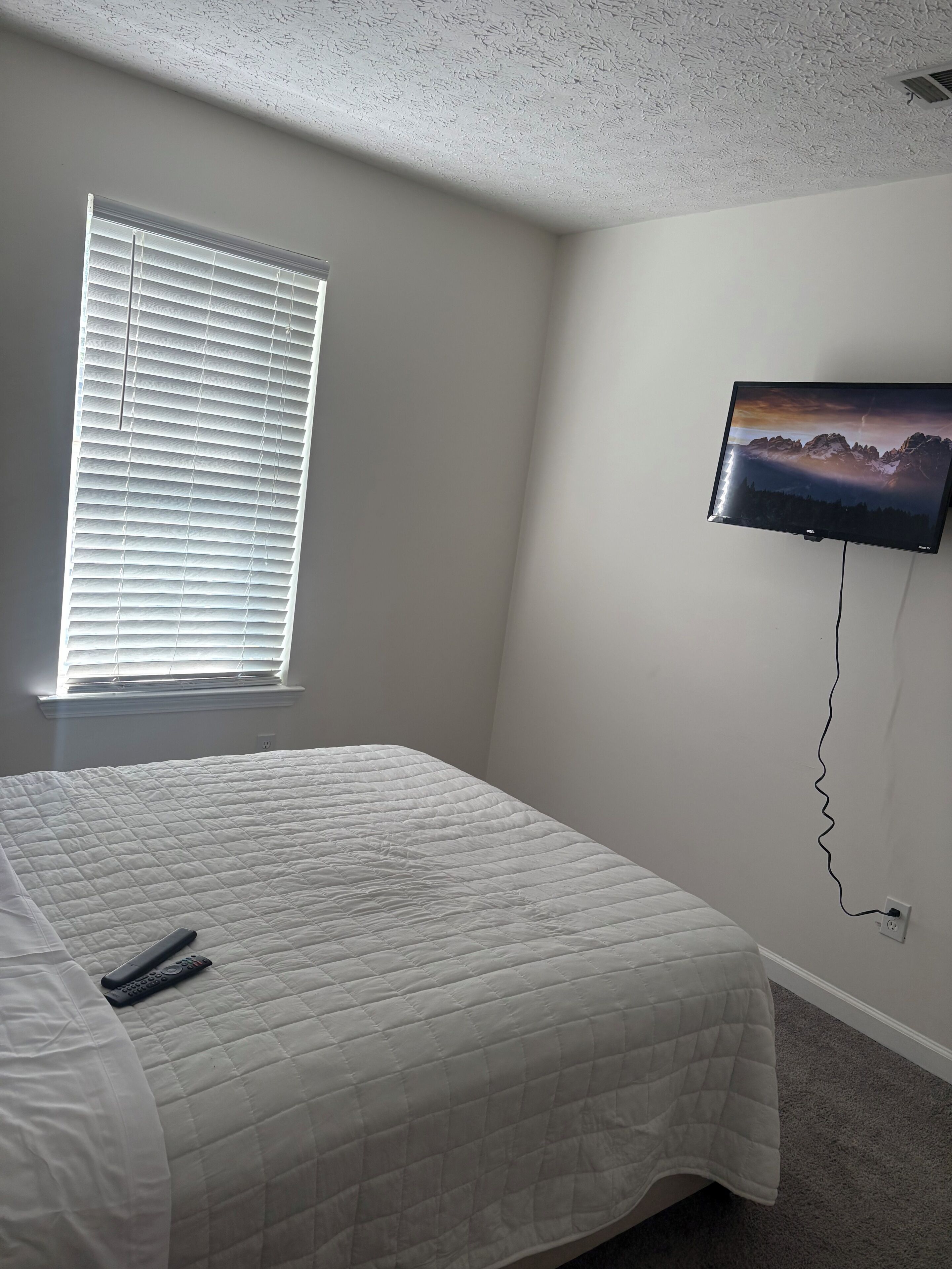 A bedroom with a bed, a television mounted on the wall, and a window with blinds.